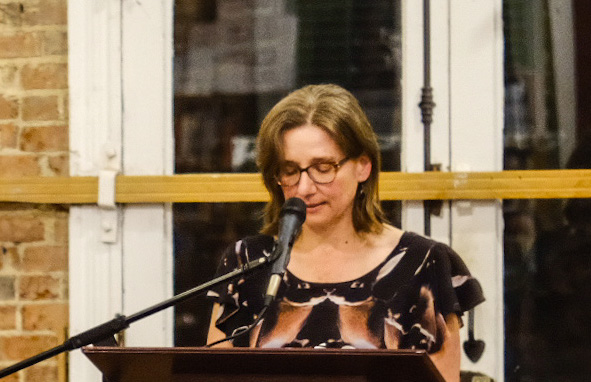 Image of Julia reading, a white woman with shoulder length reddish brown hair, glasses, and a dark shirt standing at a lectern reading into a microphone